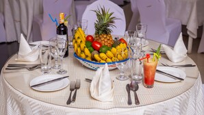 A round dining table with a white tablecloth, a bowl of fruit, a bottle of wine, and a glass of red drink.