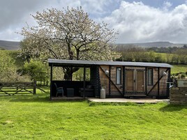 Una cabaña rústica con techo de metal, paredes de madera y cimientos de piedra. Tiene una puerta de madera, una ventana y un pequeño porche con un banco.