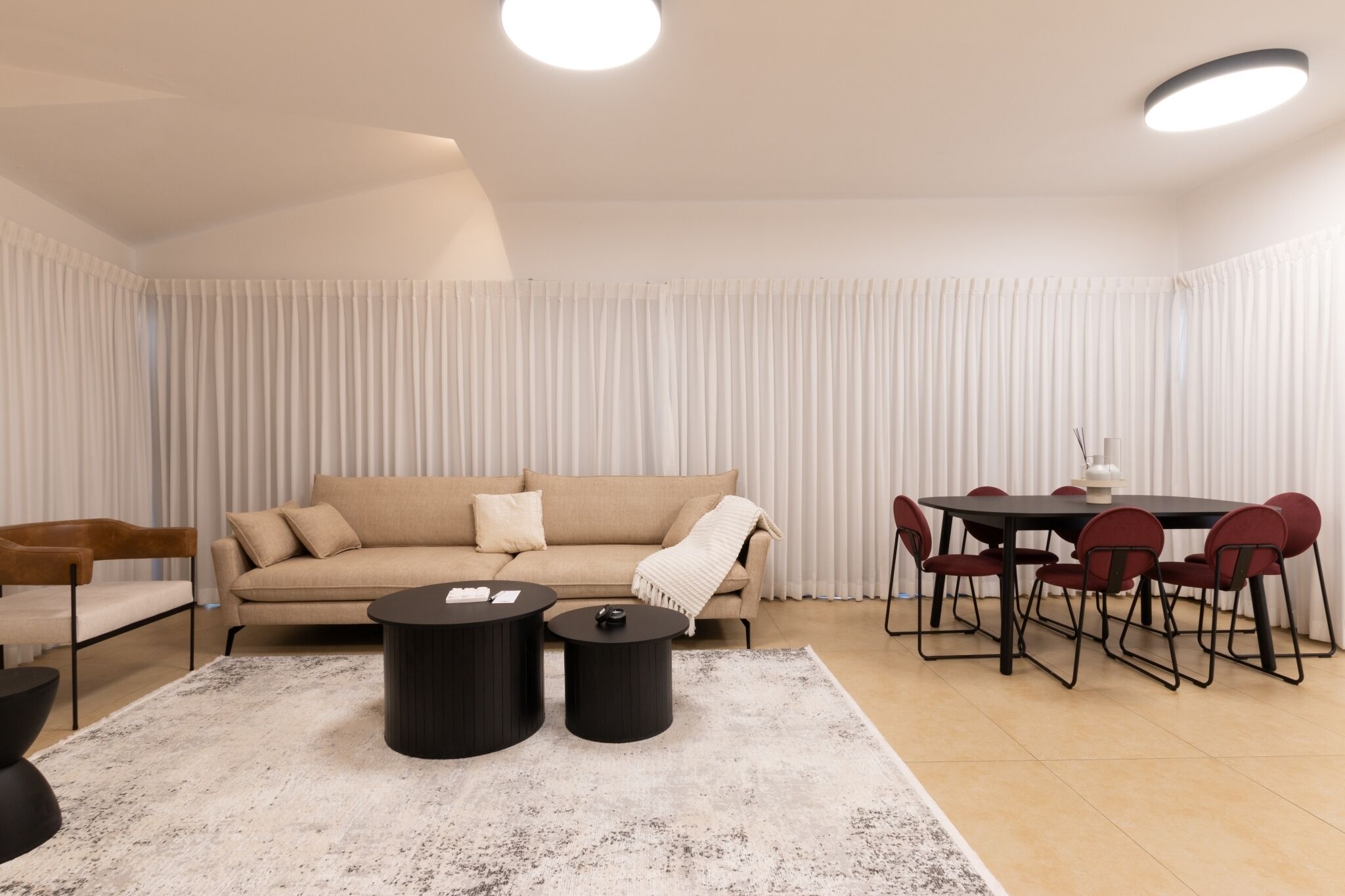 A modern living room with a beige sofa, two black side tables, a round dining table with red chairs, and a white curtain backdrop.