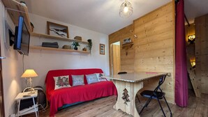 A cozy room with a red sofa, a small table, a chair, wooden walls, and a hanging light fixture. - Charming 19m² Studio for 3 with Terrace and Parking, 5min from Shops & Free Shuttle (Arêches-Beaufort)