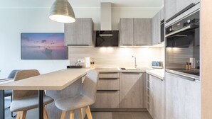 A modern kitchen with a wooden table, two grey chairs, a built-in oven, and a painting on the wall. - Ferienhaus Westwind in Tönning für bis zu 4 Personen (Tönning)