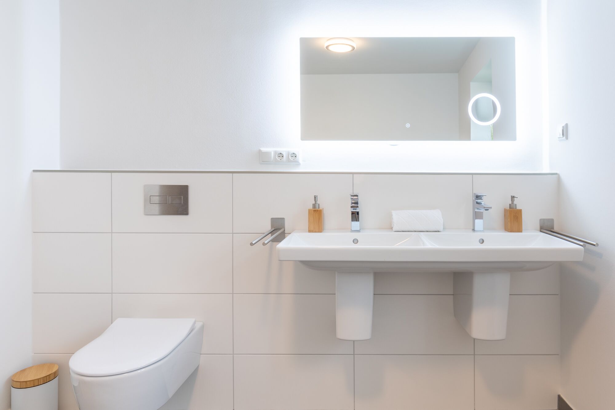 A modern bathroom with a white sink, two faucets, a mirror, and a toilet.