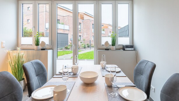 A dining area with a table set for four, large windows, and a view of a brick building and greenery outside. - Ferienhaus Westwind in Tönning für bis zu 4 Personen (Tönning)
