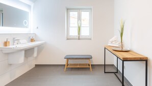 A modern bathroom with a large mirror, dual sinks, a bench, and a window with a view. - Ferienhaus Westwind in Tönning für bis zu 4 Personen (Tönning)