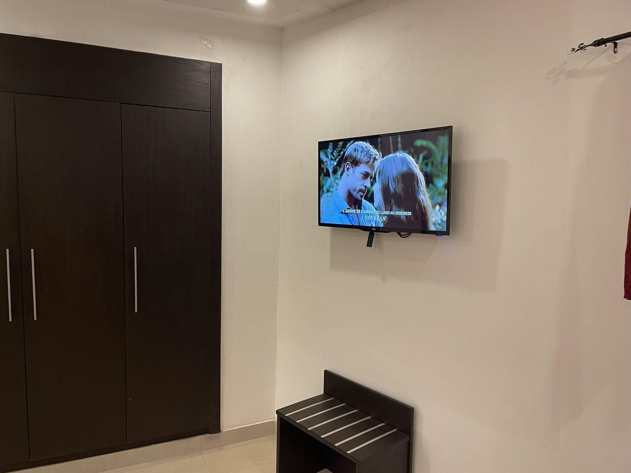 A room with a dark wardrobe, a mounted TV on the wall, and a small bench.