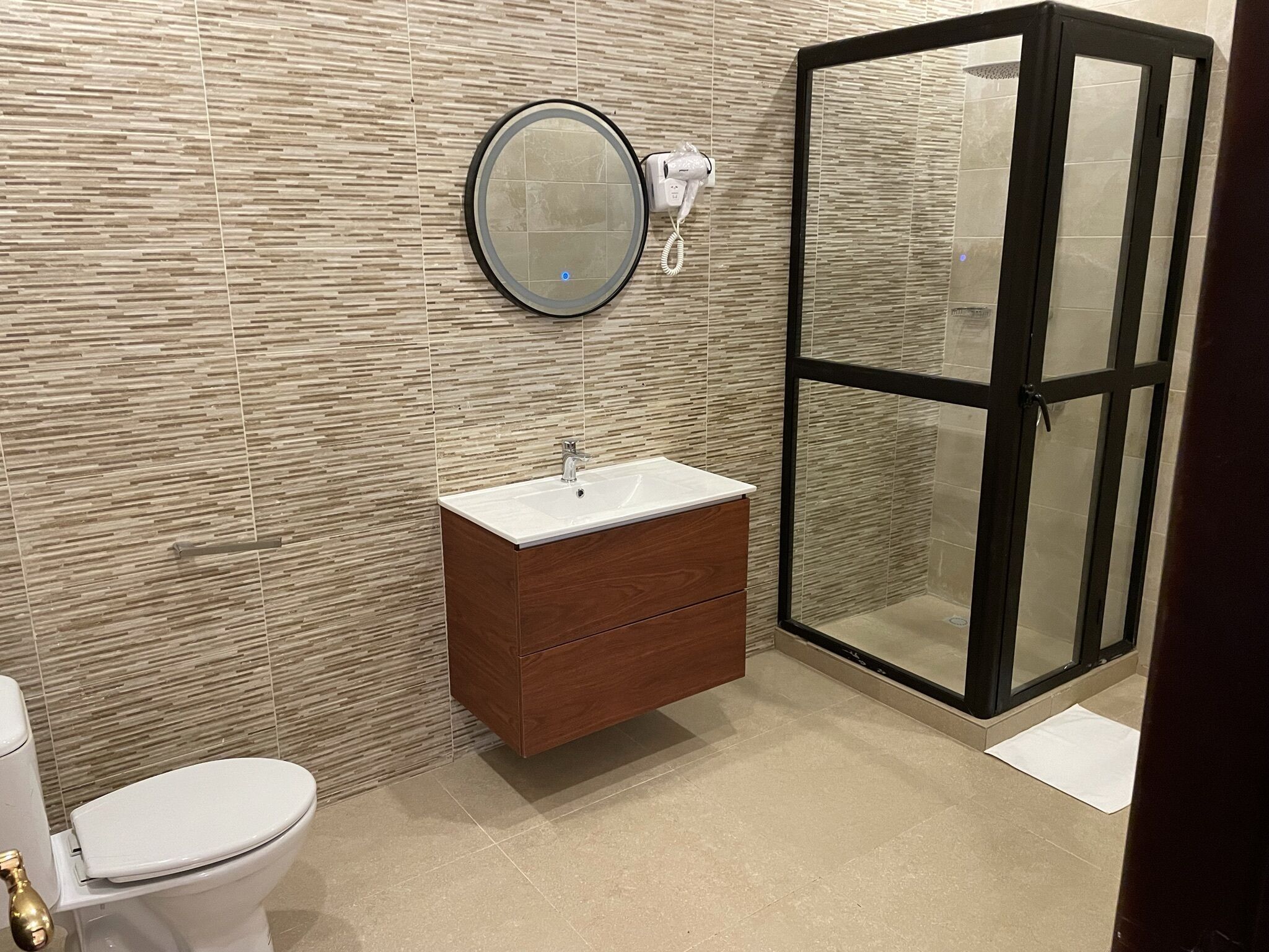 A bathroom with a toilet, a sink with a wooden vanity, a round mirror, and a glass-enclosed shower.