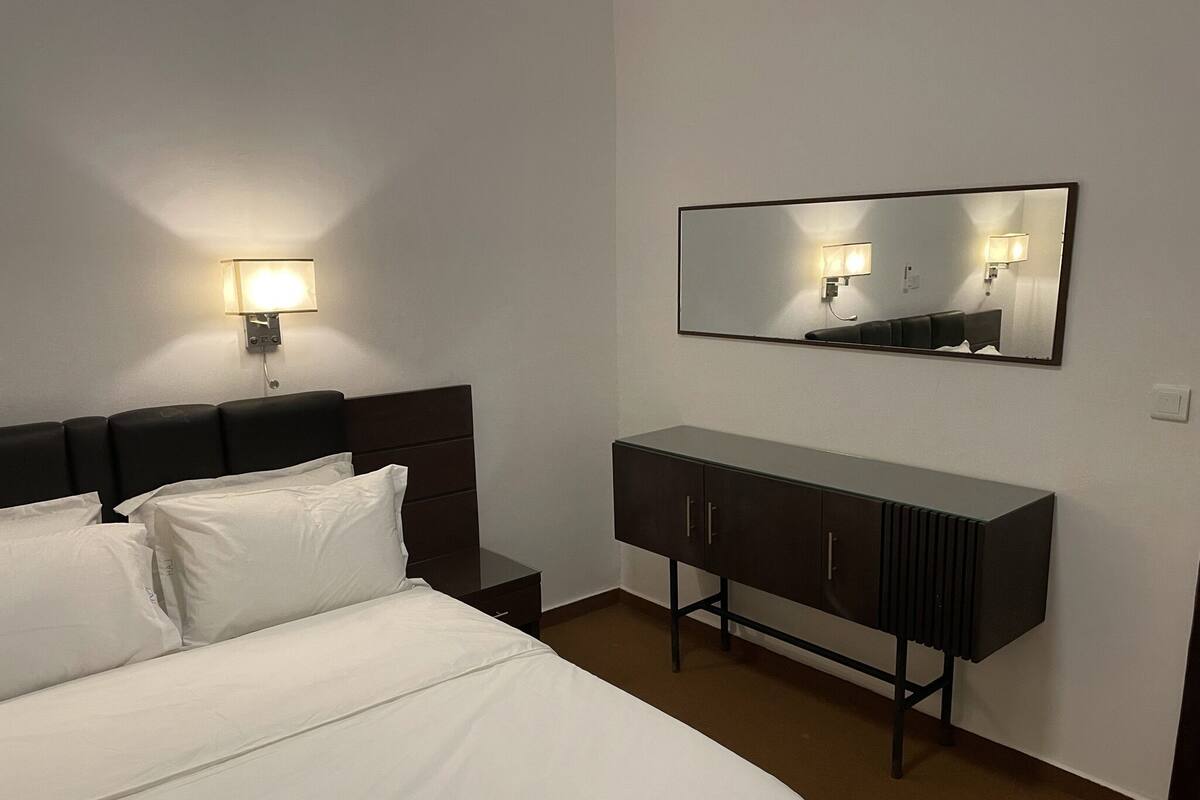 Habitació d'hotel amb llit, tauleta de nit, prestatge i mirall fixat a la paret.