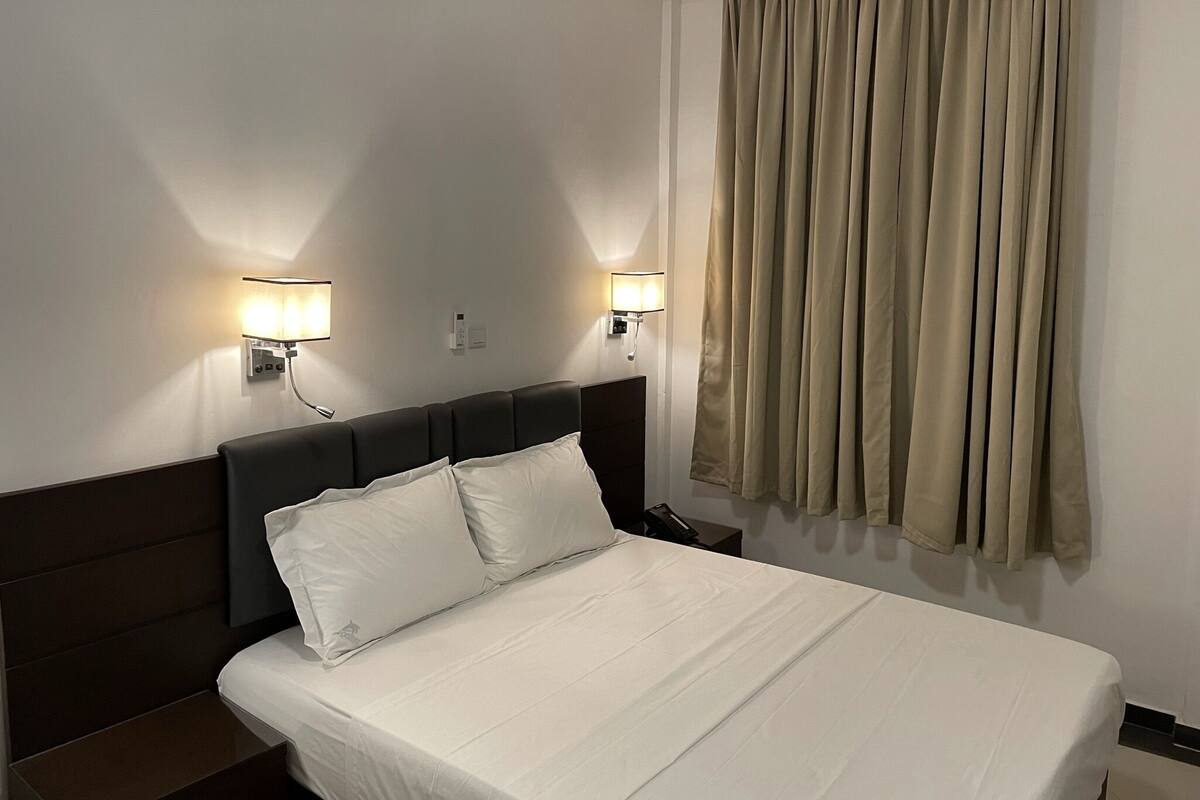Habitació d'hotel amb un llit, dues làmpades de nit, una finestra amb cortines i un interruptor fixat a la paret.