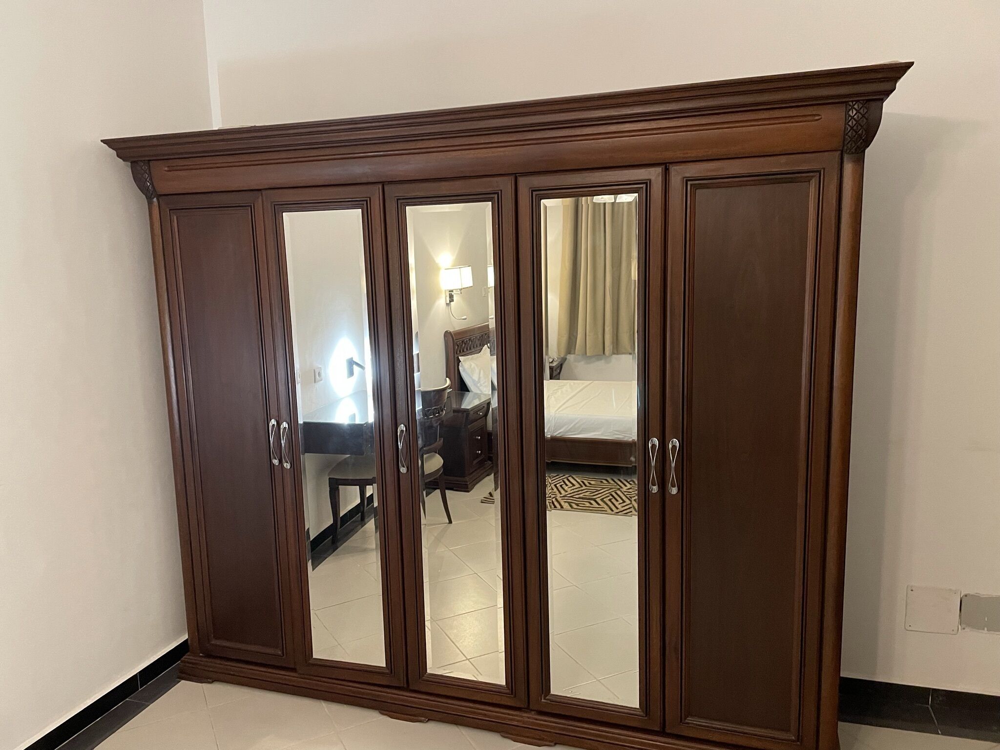 A large wooden wardrobe with mirrored doors, a bed with a white mattress and beige curtains, and a small desk with a lamp.