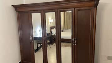A large wooden wardrobe with mirrored doors, a bed with a white mattress and beige curtains, and a small desk with a lamp.