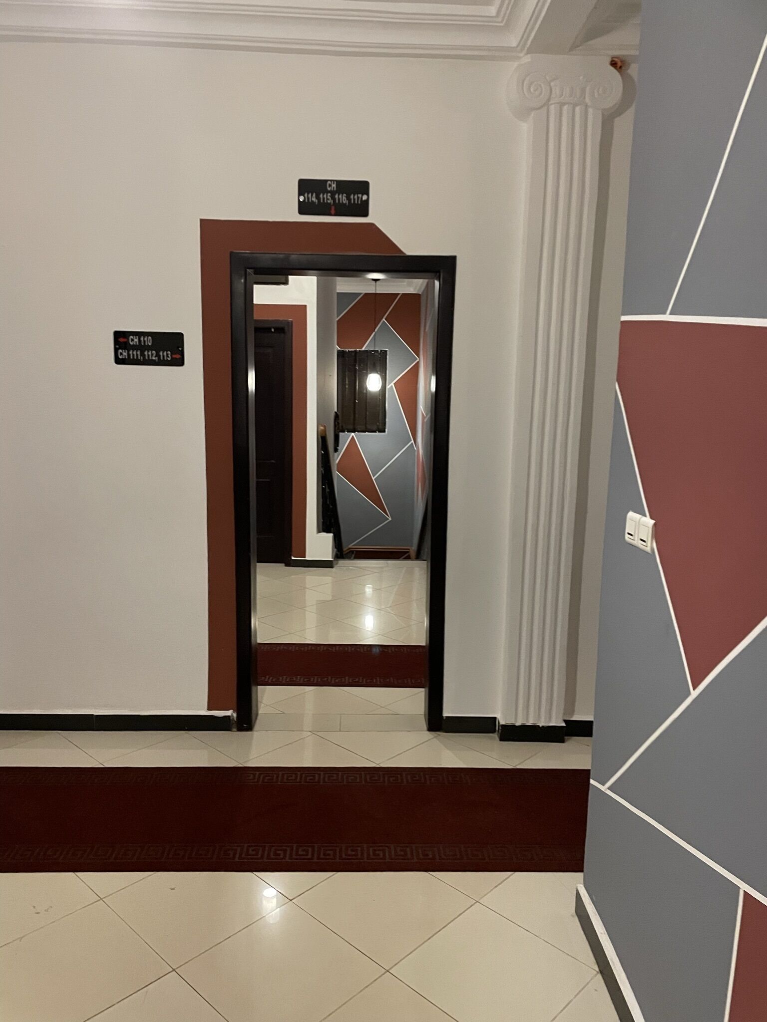 A hallway with a geometric-patterned wall, a door with a sign indicating room numbers, and a tiled floor.