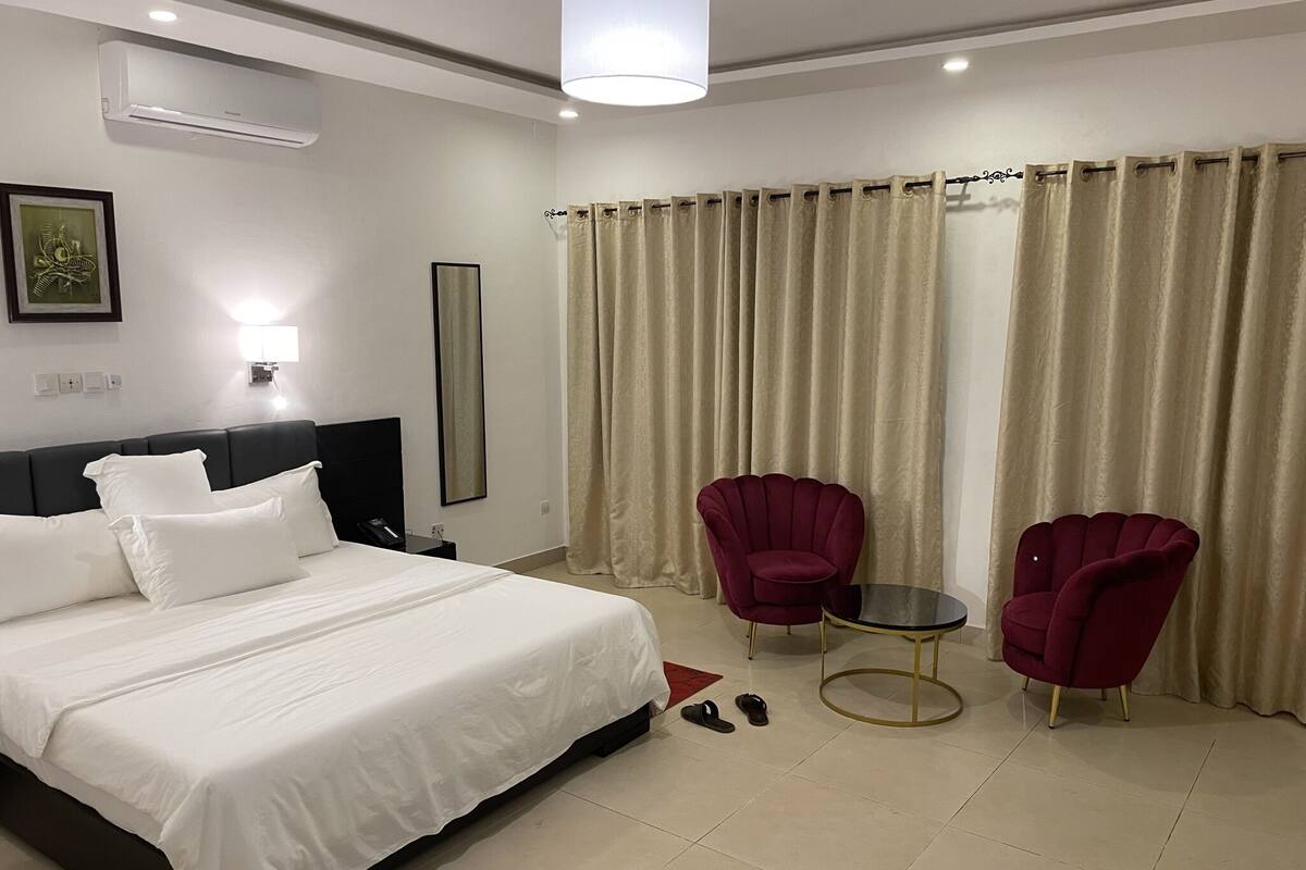 Habitació d'hotel amb un llit, dues butaques, una taula petita i una finestra amb cortines.