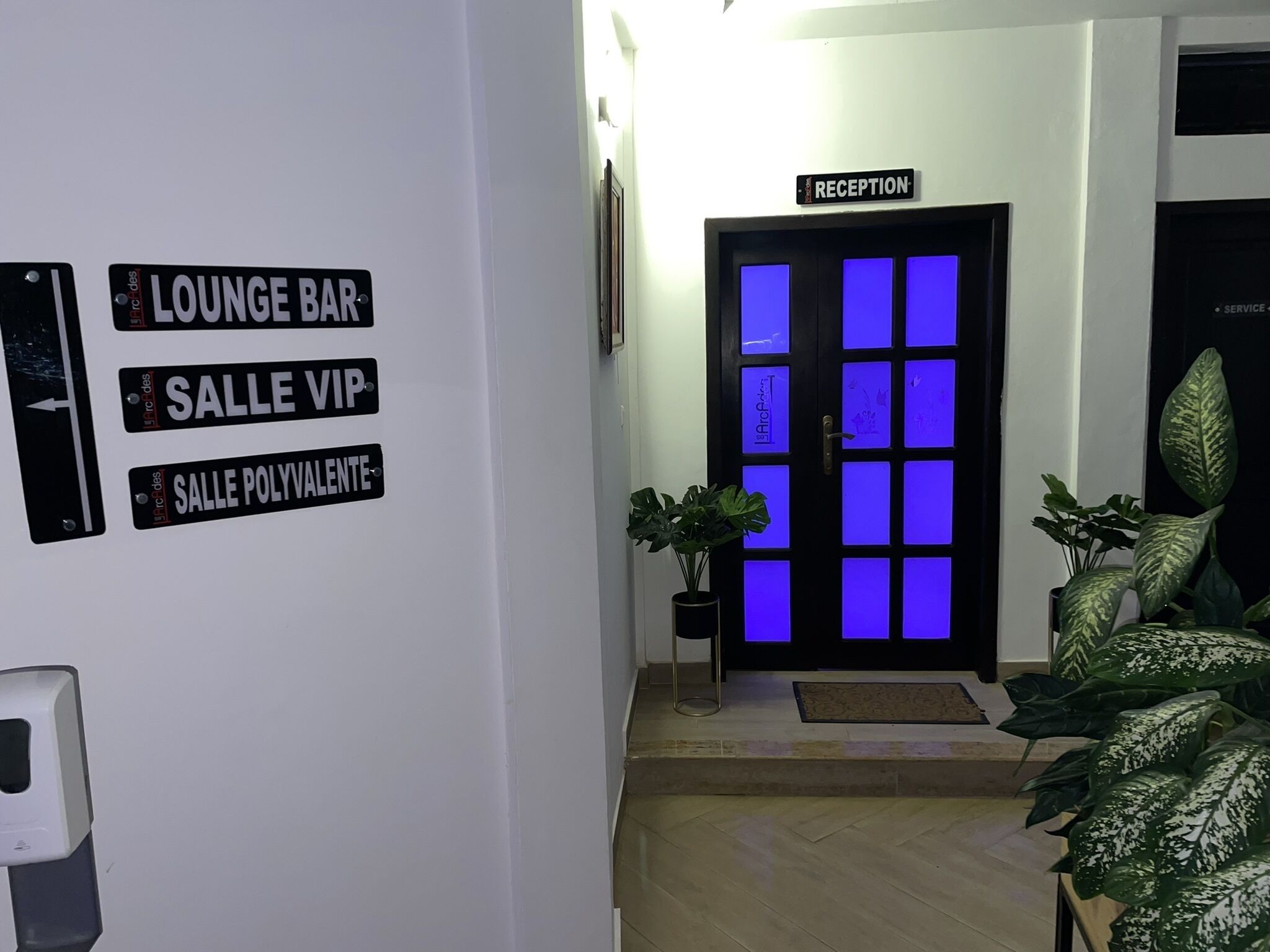 Un área de recepción con un cartel que indica 'RECEPTION' sobre una puerta. Hay letreros que dicen 'LOUNGE BAR', 'SALLE VIP' y 'SALLE POLYVALENTE' en la pared.