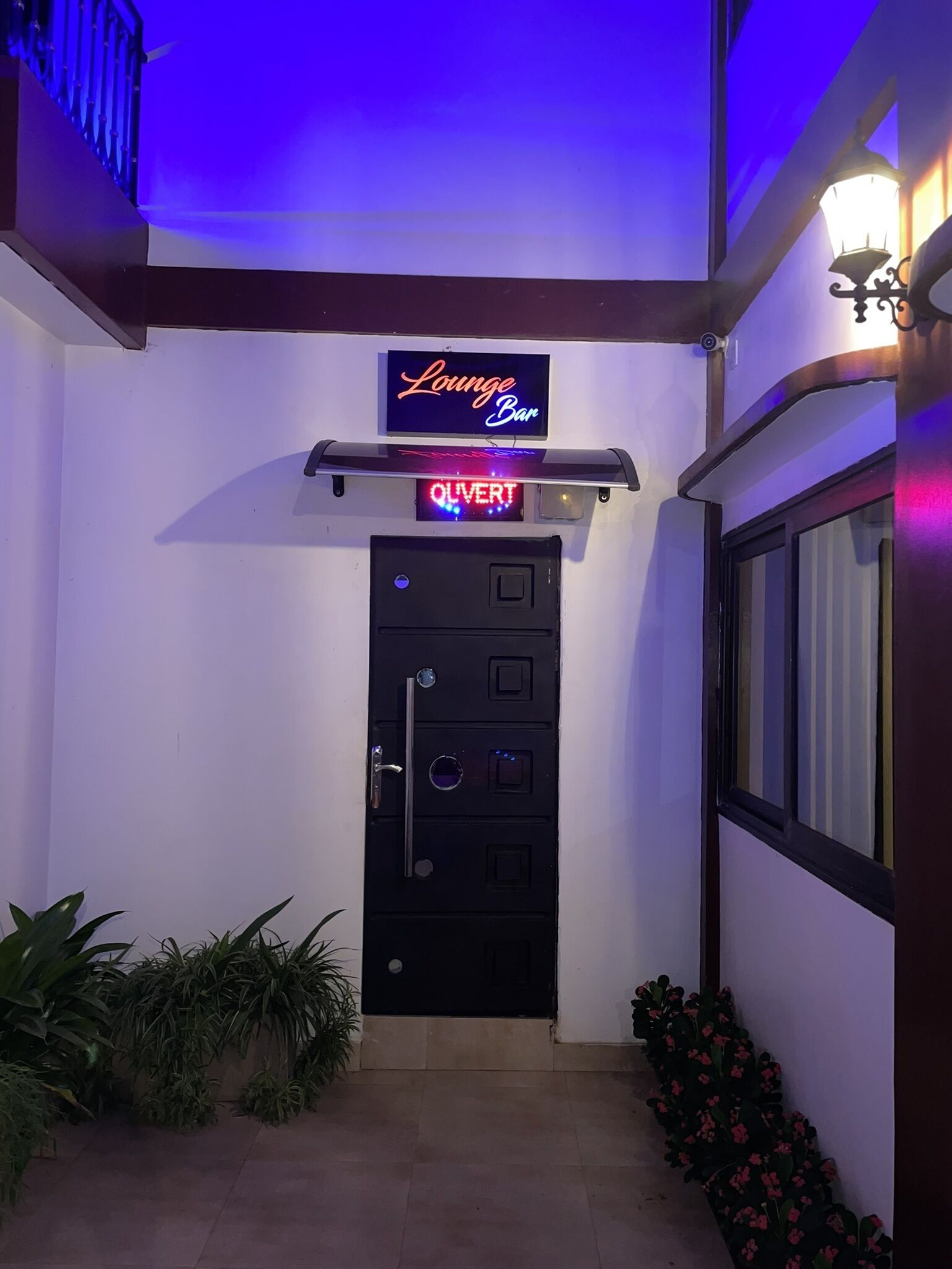 A black door with a digital sign above it, a neon sign reading "Lounge Bar", and a security keypad.