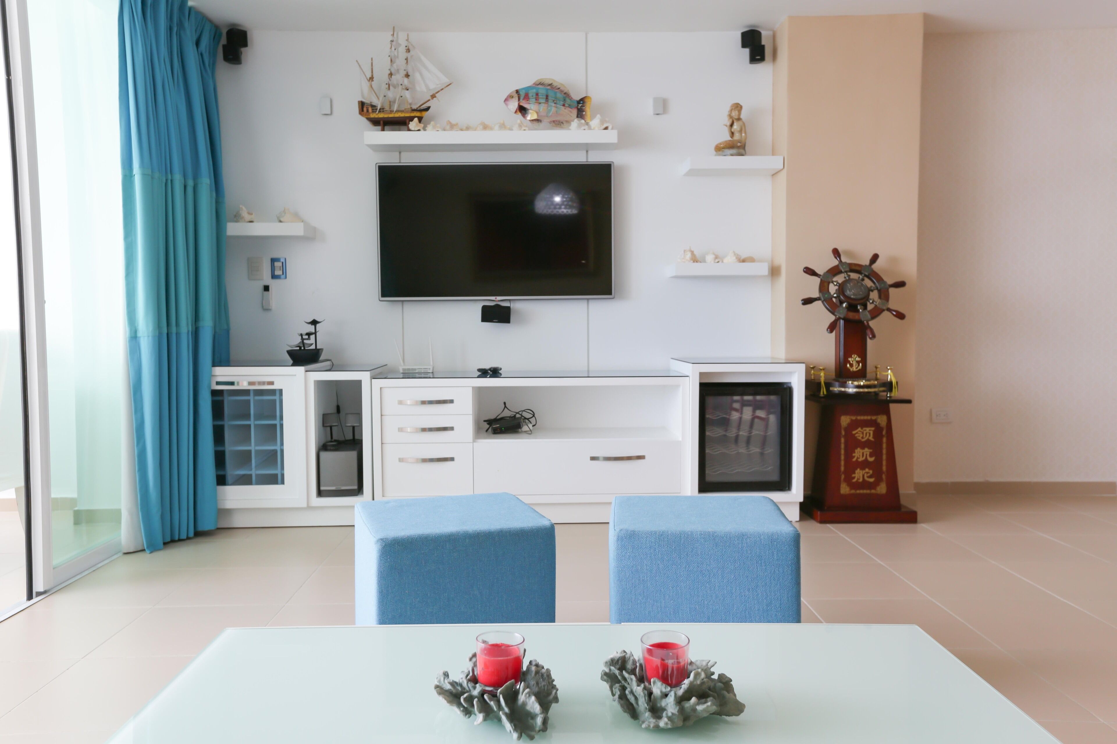 A modern living room with a flat-screen TV, blue upholstered chairs, a small table with candles, and nautical decorations.
