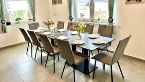 A dining area with a table set for six, surrounded by chairs, with a view of the outdoors through large windows.