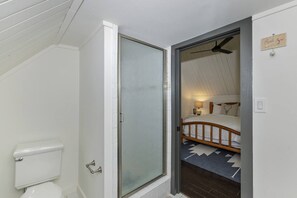 A bathroom with a toilet, a glass shower door, and a bed in the adjacent room.