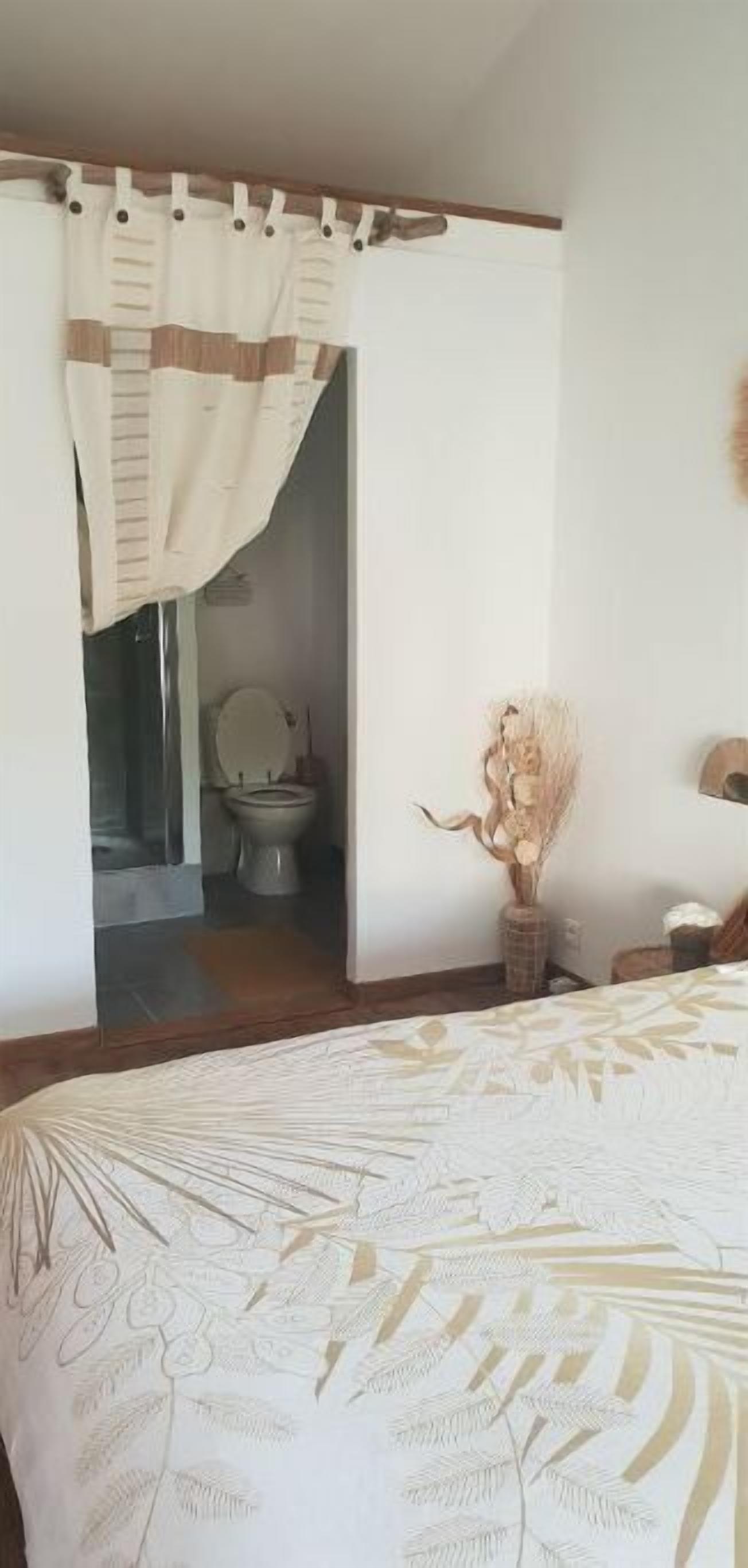 Une chambre à coucher avec un lit, une salle de bain visible à travers une porte ouverte, et un vase contenant des fleurs séchées sur une table de chevet.