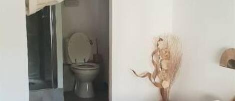 Une chambre à coucher avec un lit, une salle de bain visible à travers une porte ouverte, et un vase contenant des fleurs séchées sur une table de chevet.