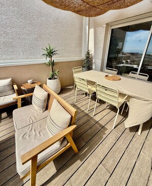 Un espace extérieur ensoleillé, aménagé avec une terrasse en bois, des fauteuils confortables, une table ronde et des plantes en pot.