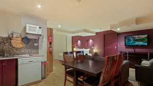 A modern kitchen with a microwave, a sink, and a dish rack. There is a dining area with a table and chairs. A television is mounted on the wall.