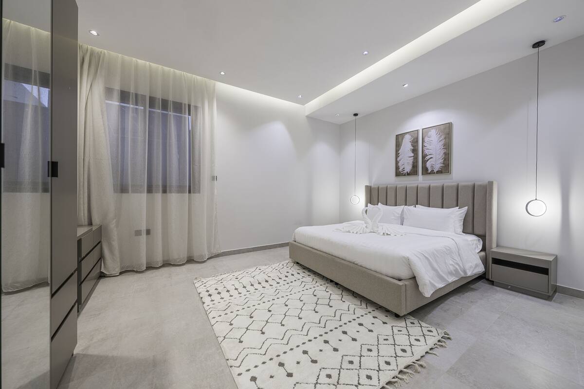 A modern bedroom with a large bed, bedside tables, a patterned rug, and a ceiling light fixture.