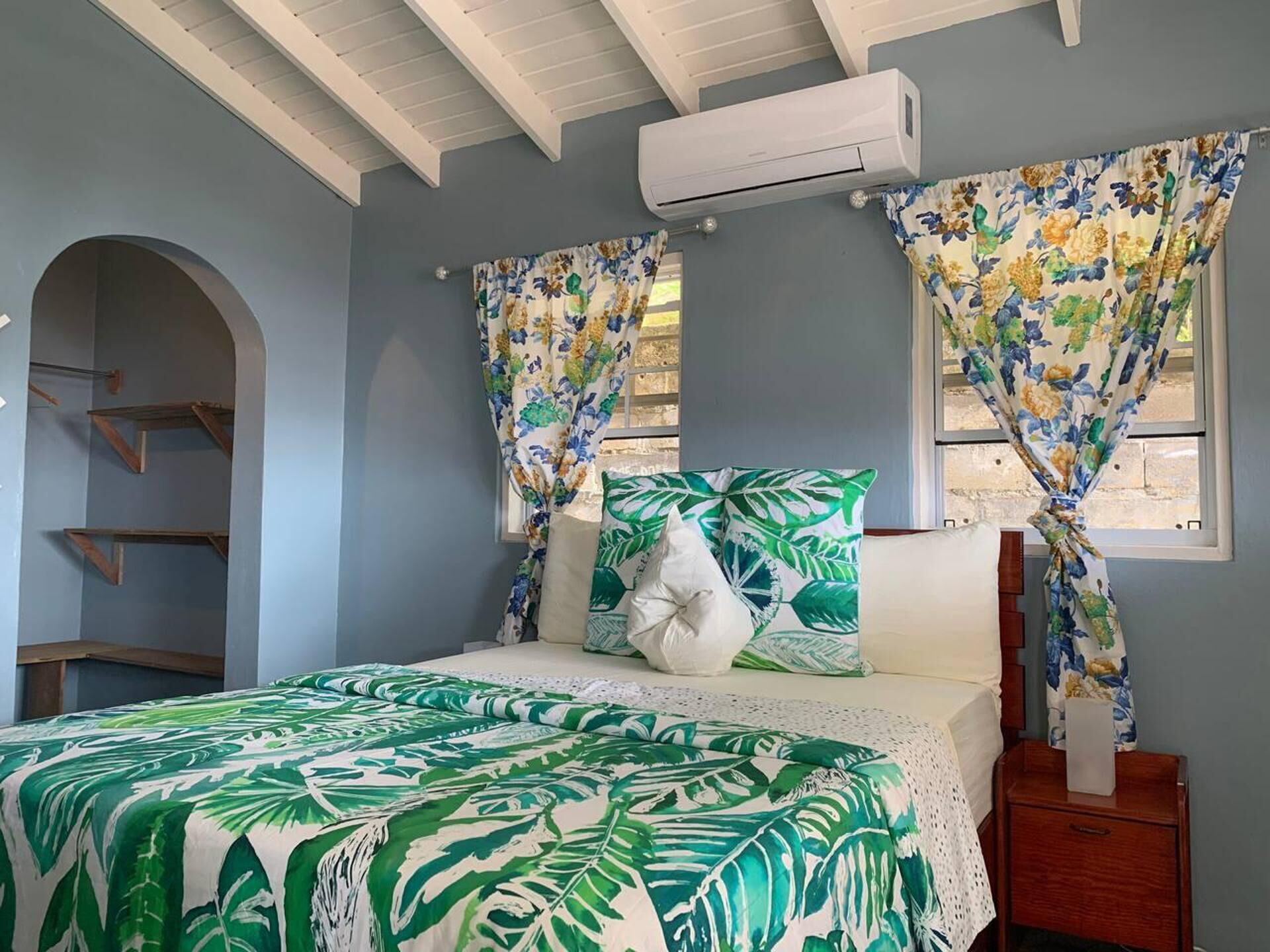 A bedroom with a bed, floral curtains, a white air conditioning unit, and a wooden nightstand.