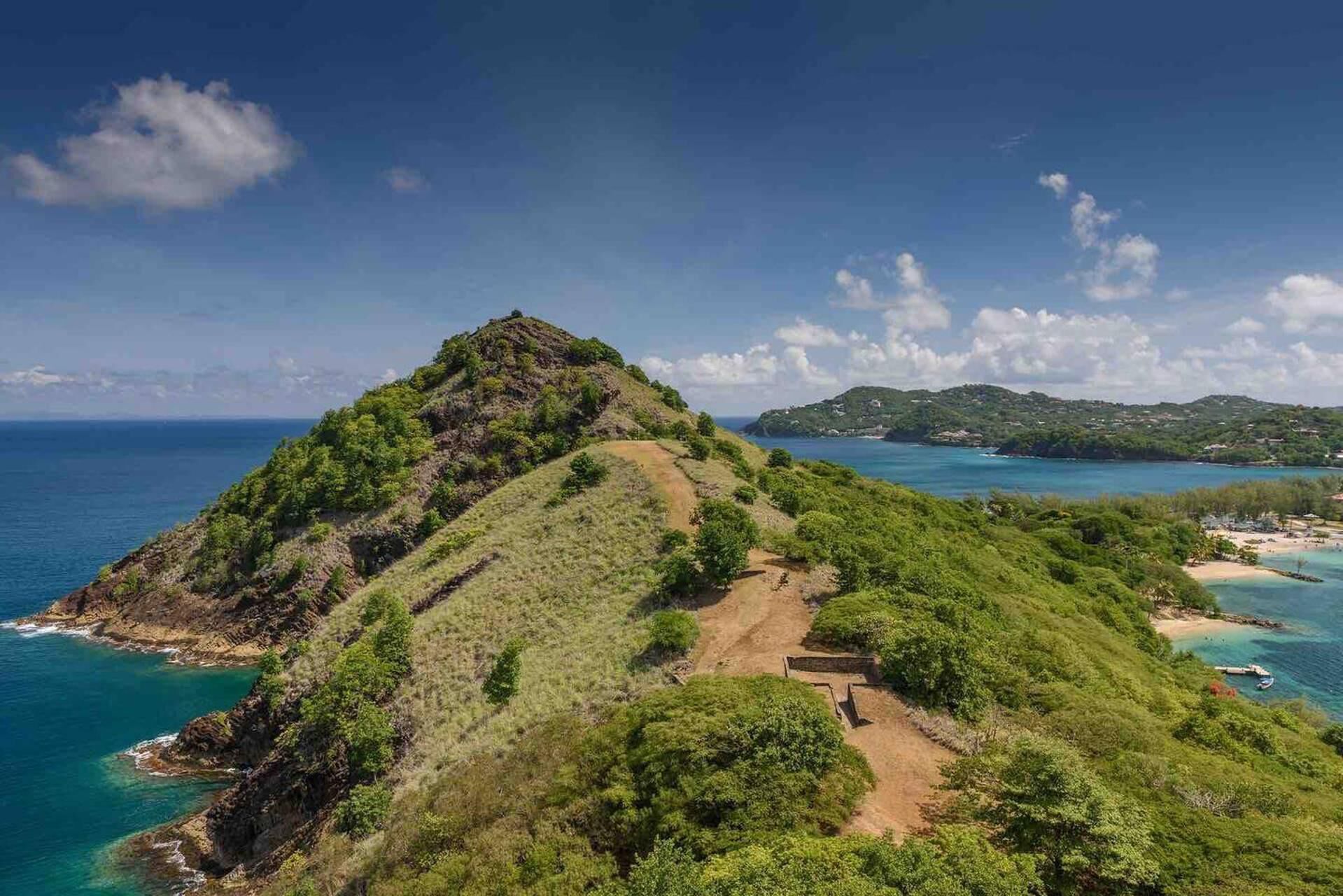 Una colina exuberante y verde con un sendero de tierra que lleva a una estructura, con vistas a una zona costera de aguas azules cristalinas y islas distantes.