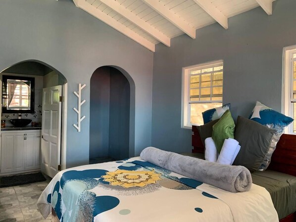 A bedroom with a bed, pillows, a window, and a kitchen area visible through an archway.
