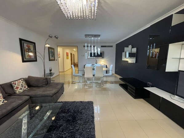 Un salon moderne avec un canapé, une table basse en verre et un tapis noir. On y trouve également un coin repas avec une table et des chaises blanches.