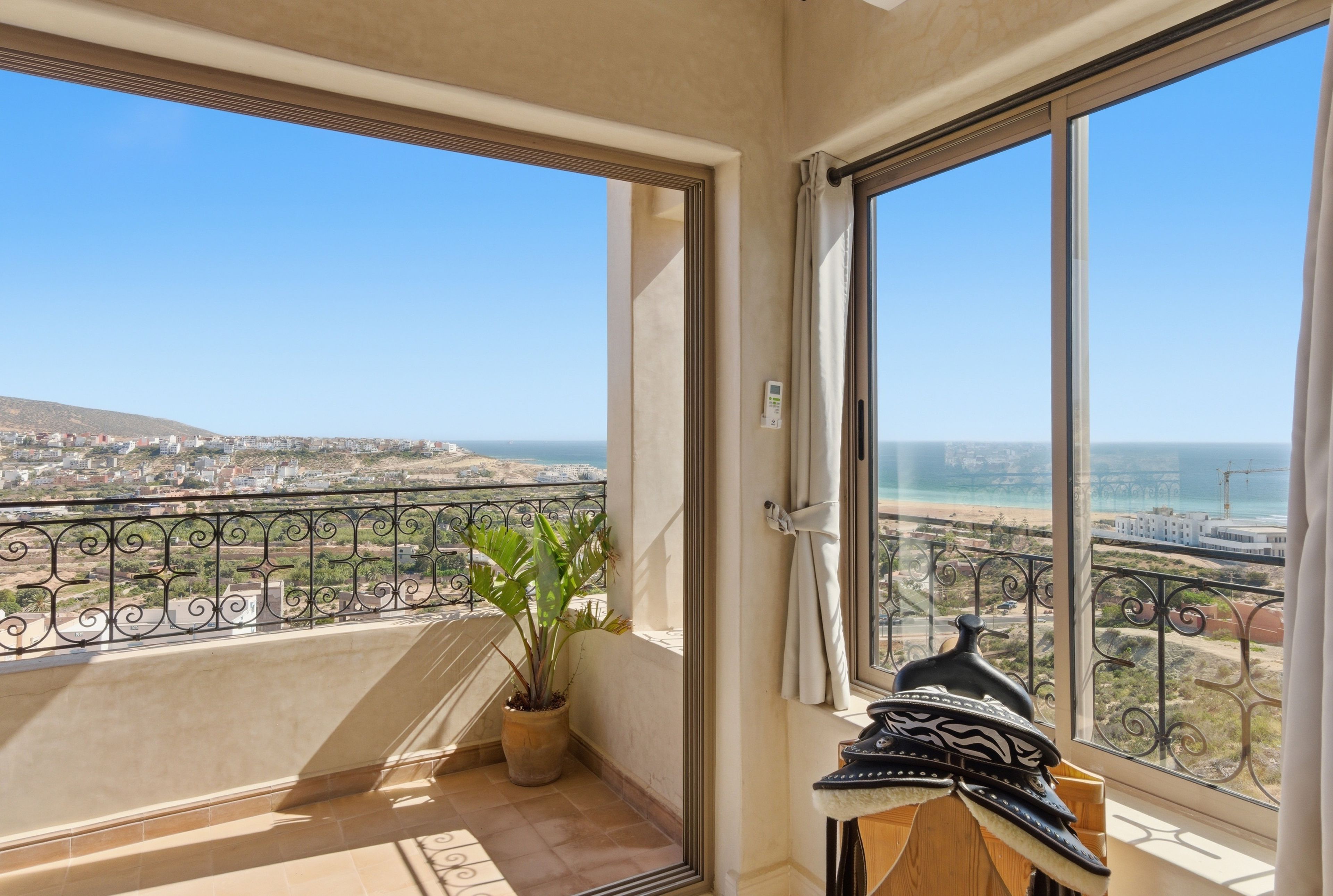 A balcony with a potted plant, a view of a coastal cityscape, and a beach with construction cranes.