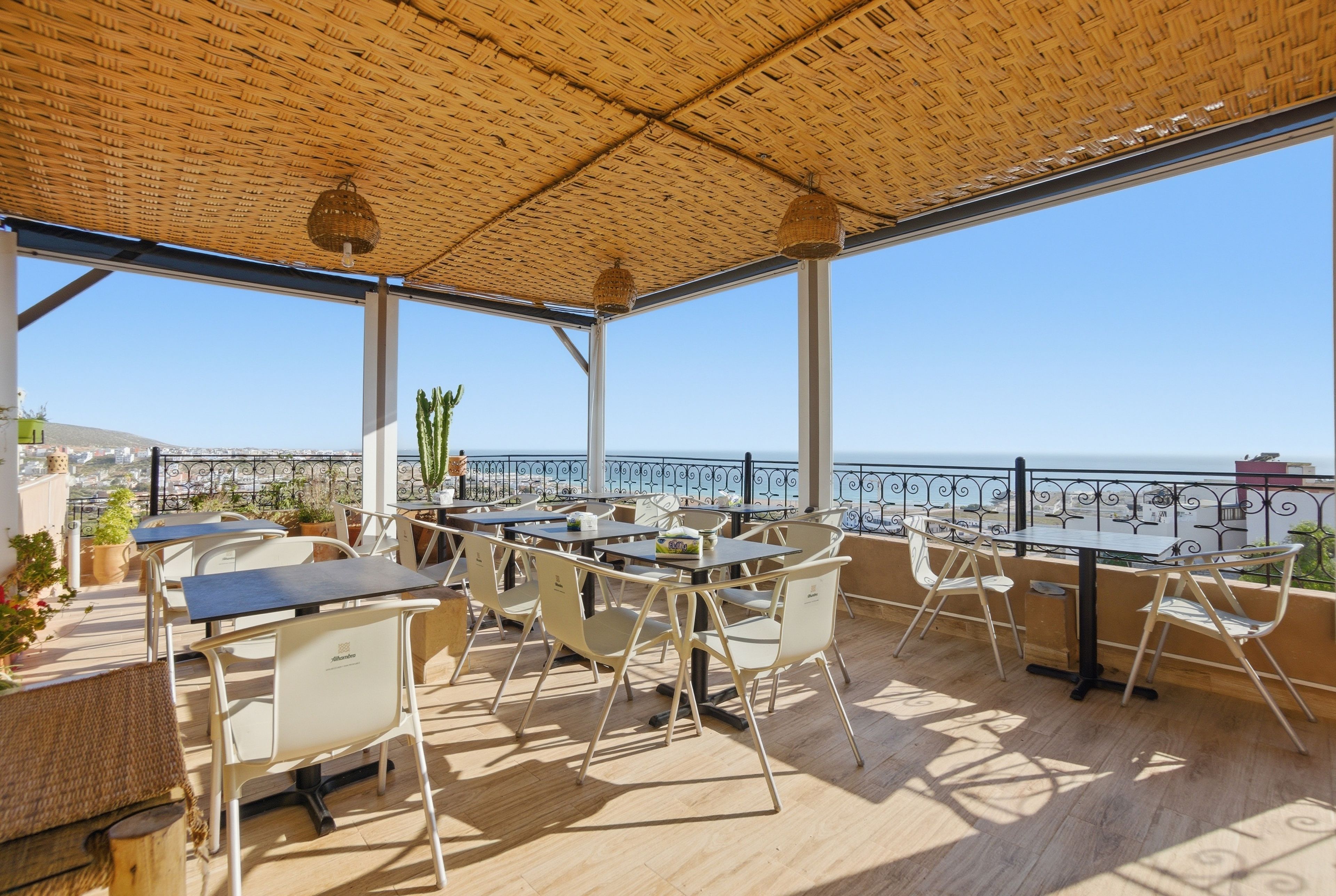 An outdoor terrace with tables and chairs, a view of the sea, and a thatched roof.