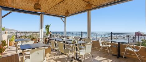 An outdoor terrace with tables and chairs, a view of the sea, and a thatched roof.