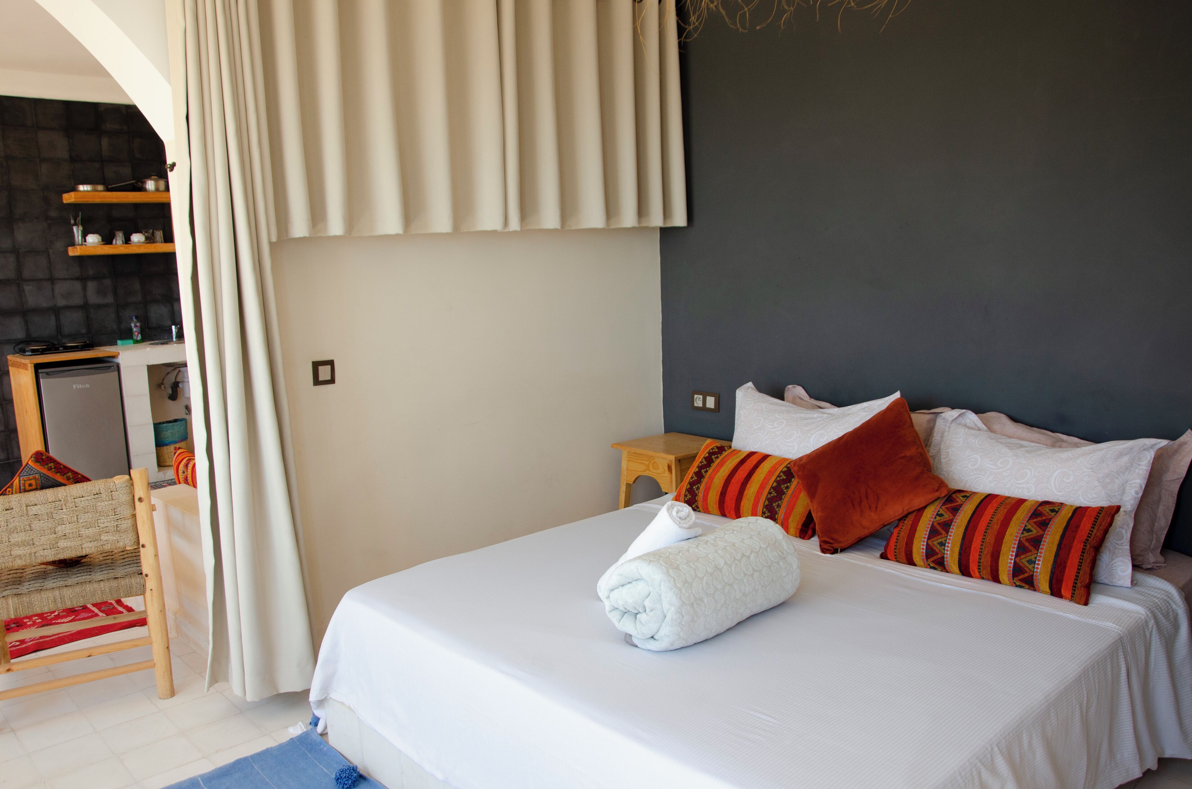 A neatly made bed with a rolled towel, a wooden nightstand with a lamp, and a small kitchenette area with shelves.
