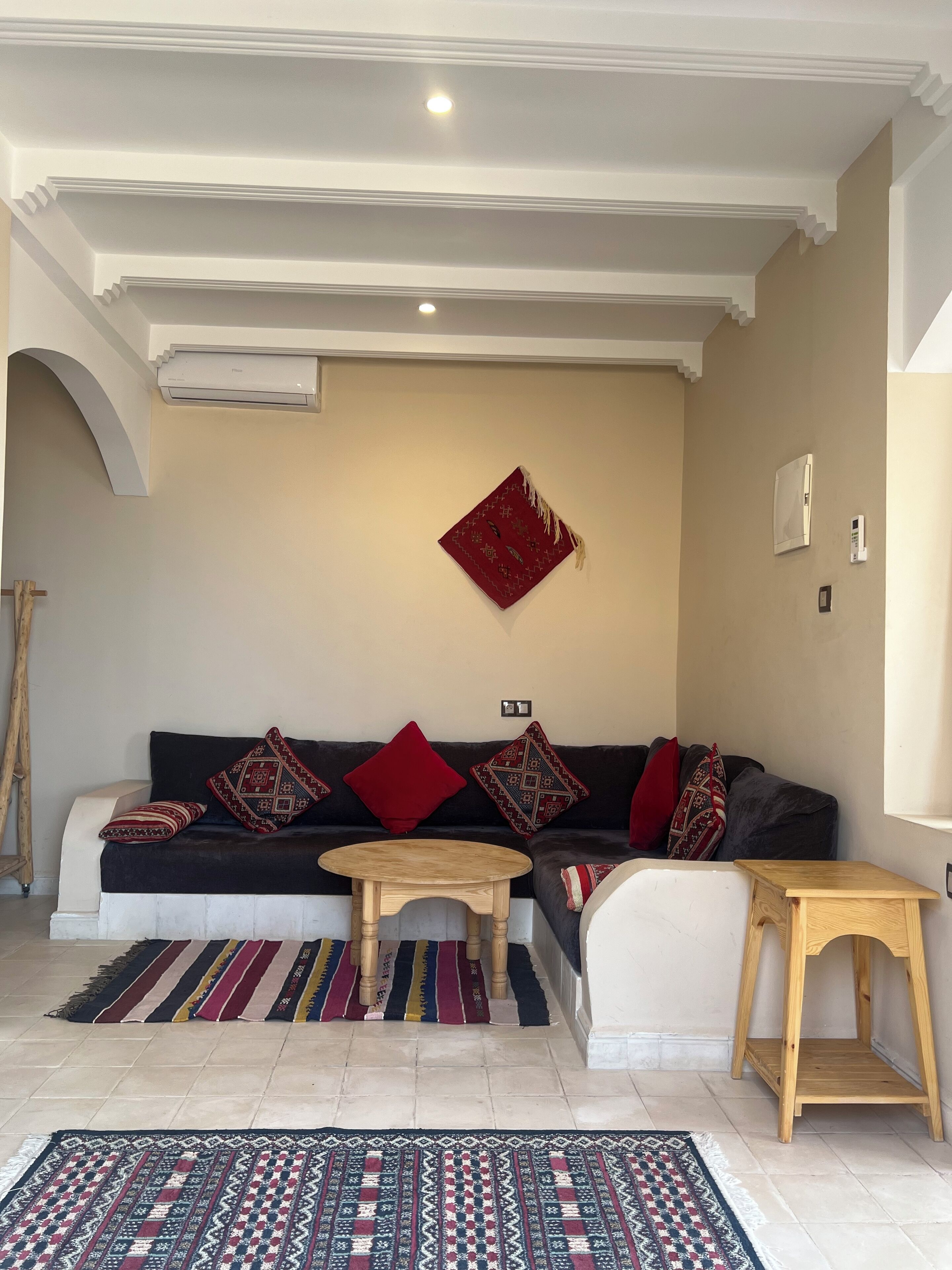 A living room with a dark-colored sofa, red cushions, a patterned rug, a small wooden table, and a wall-mounted air conditioning unit.