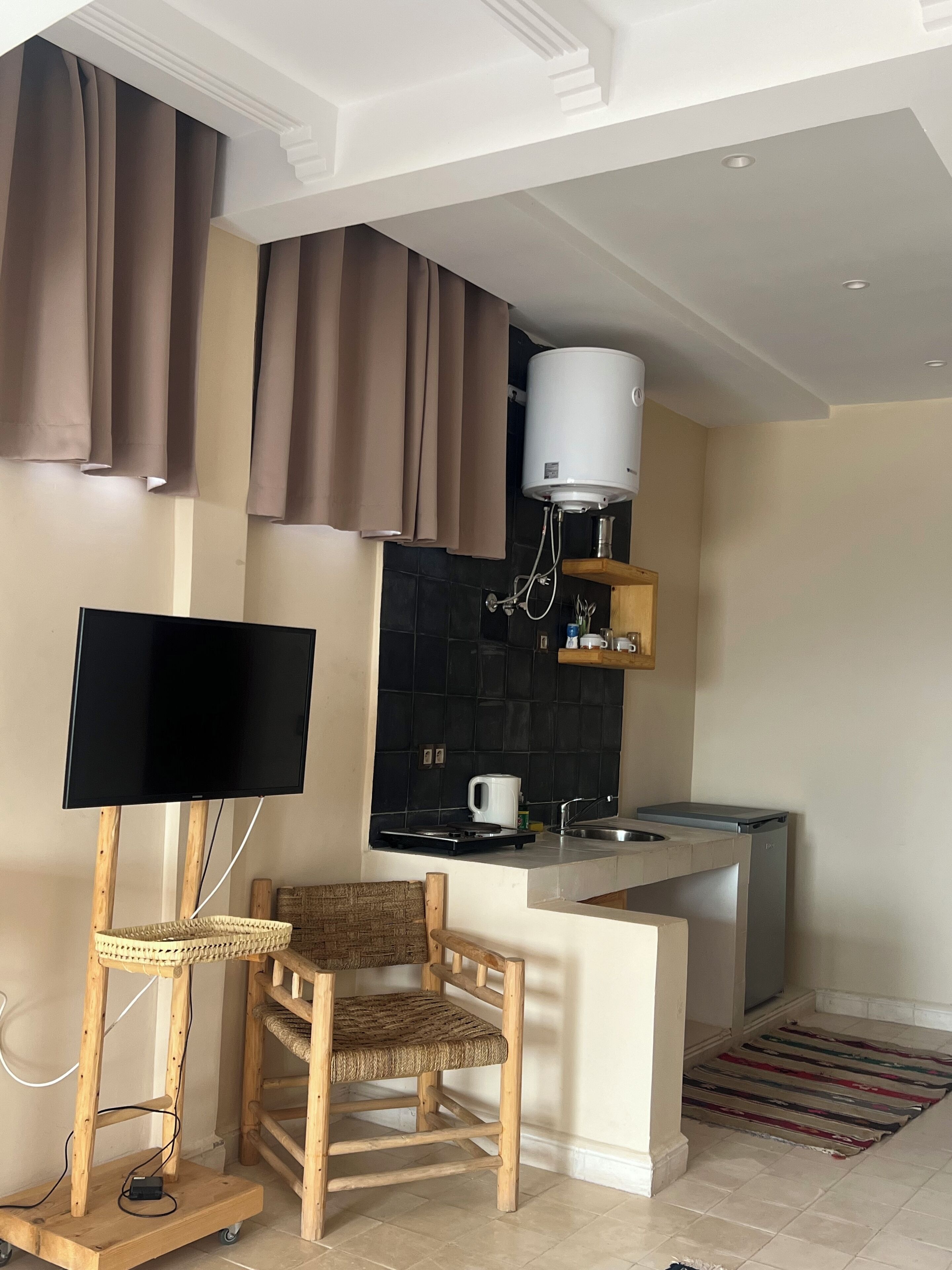 Cocina compacta con televisor de pantalla plana, calentador de agua, cafetera y una pequeña zona de comedor con una silla.