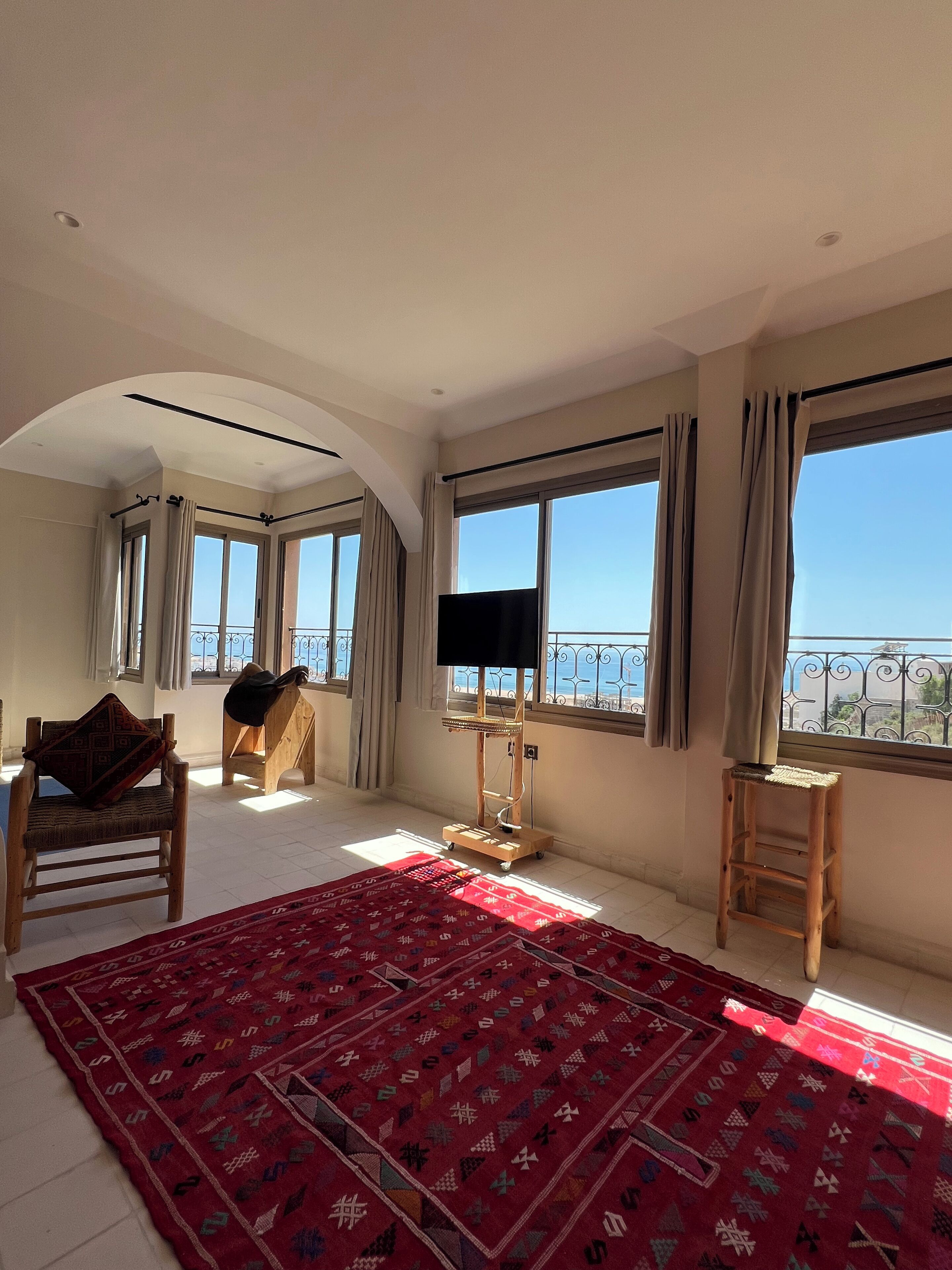 Amplia habitación con ventanas grandes, un tapete rojo estampado, una silla de madera, un taburete pequeño de madera y un mueble para televisión.