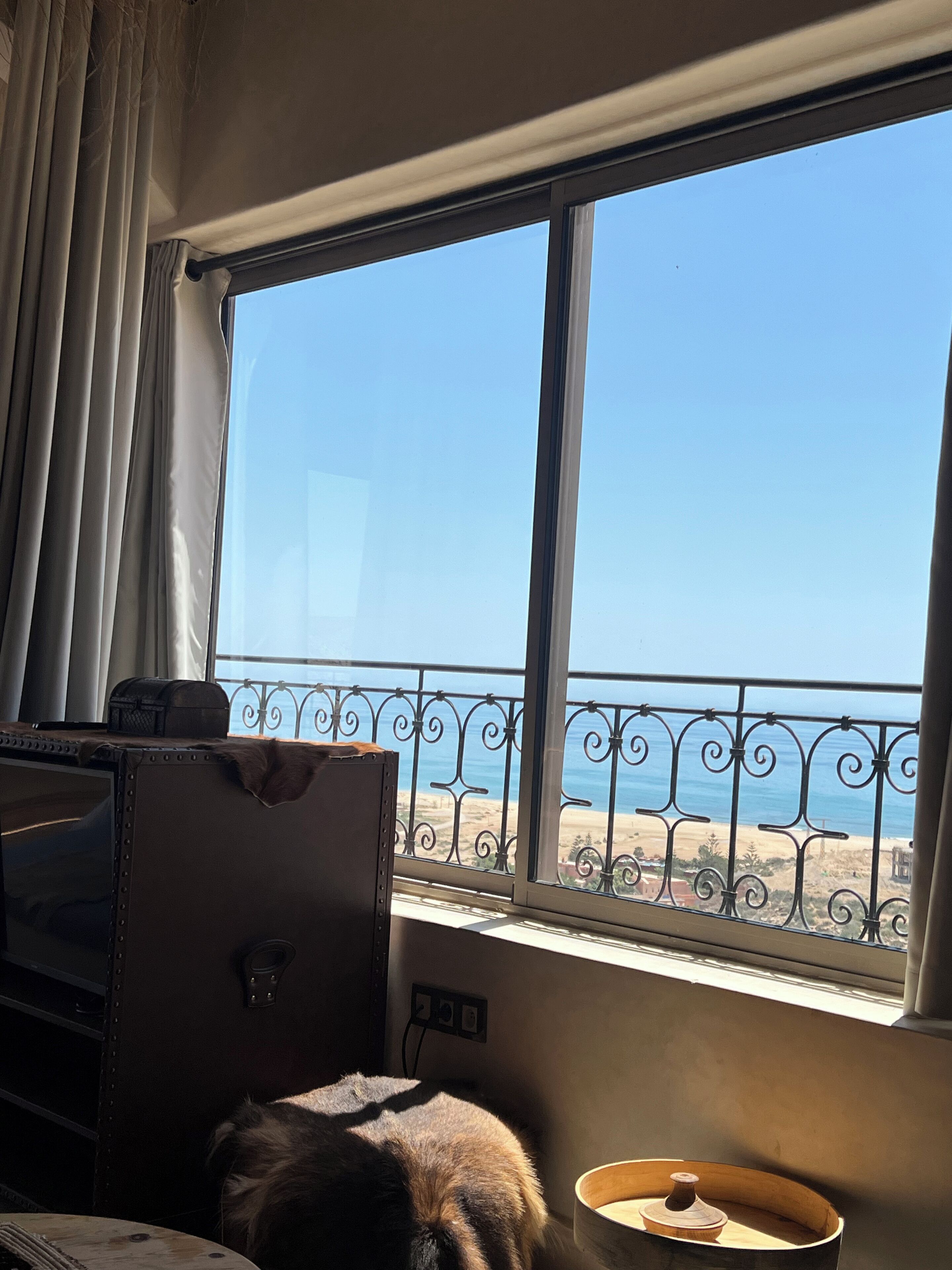 A room with a large window overlooking a beach, a suitcase, a small table with a bowl, and a power outlet on the wall.