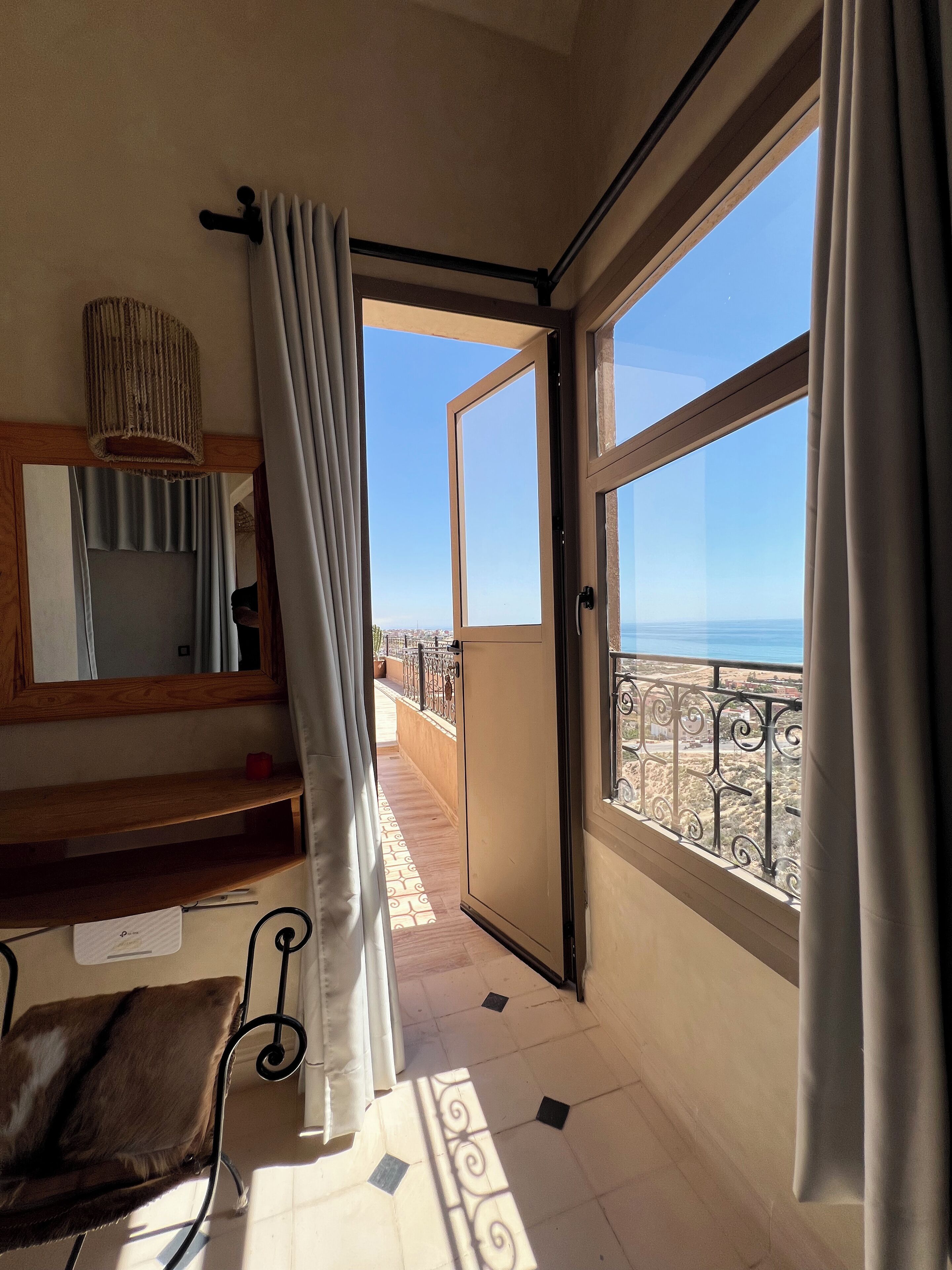 A balcony with a view of the sea, a wooden bench, a chair, a mirror, and a curtain.