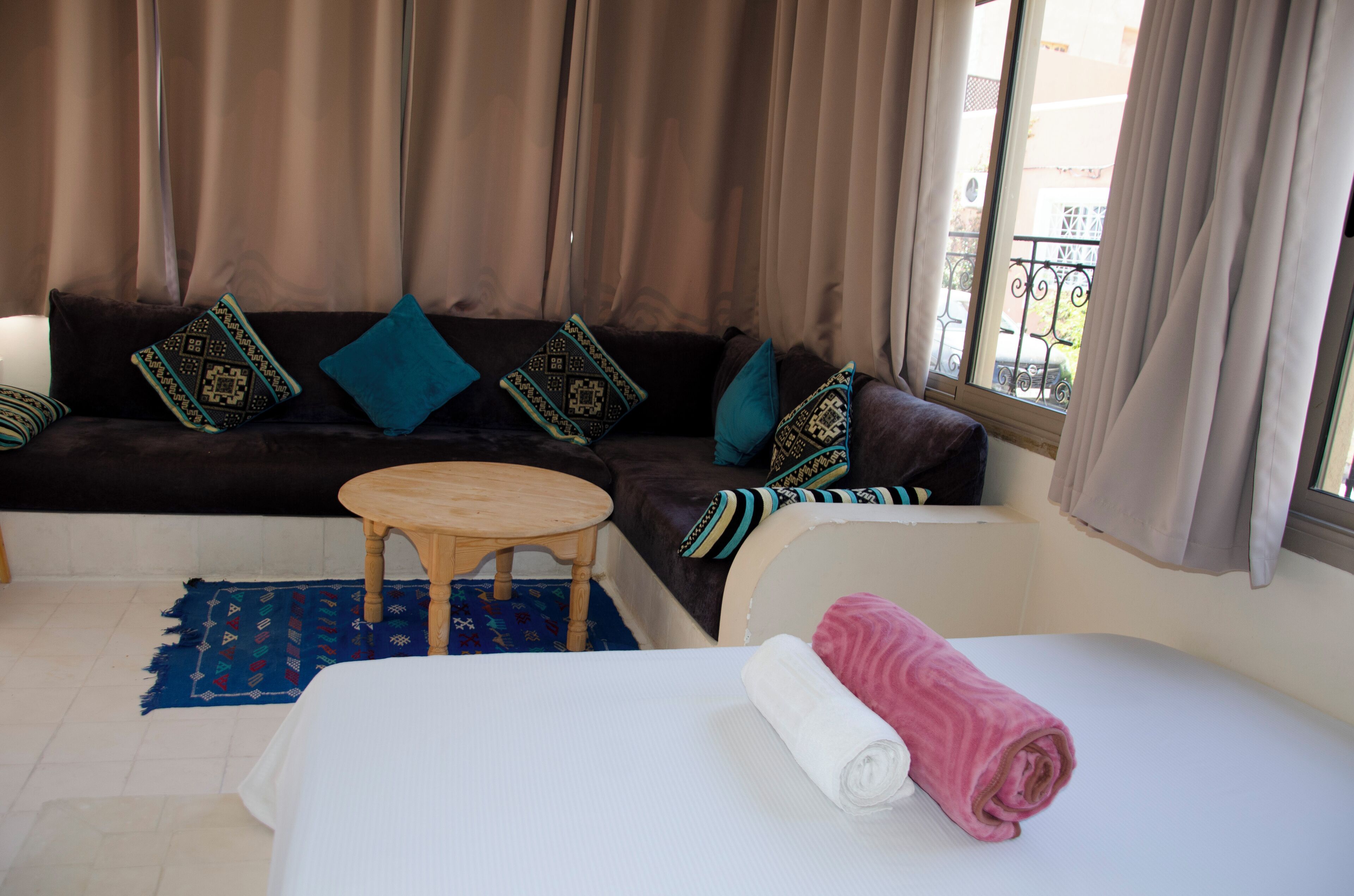 A room with a sofa, a small round table, a bed with a rolled-up pink towel, and a blue patterned rug.