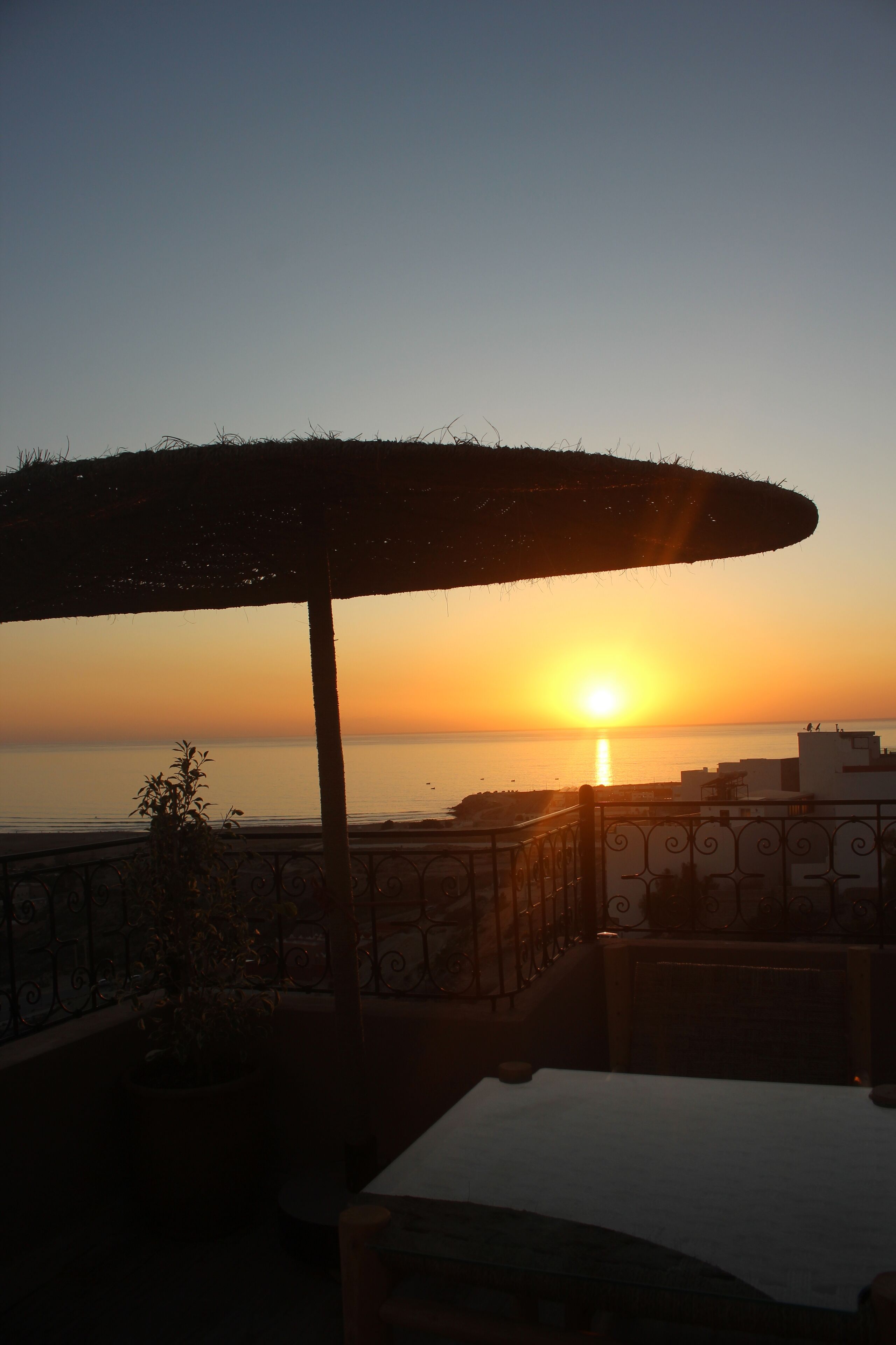 Ein Sonnenuntergangsblick von einer Terrasse mit Tisch und Stühlen, mit Blick auf das Meer und Gebäude.