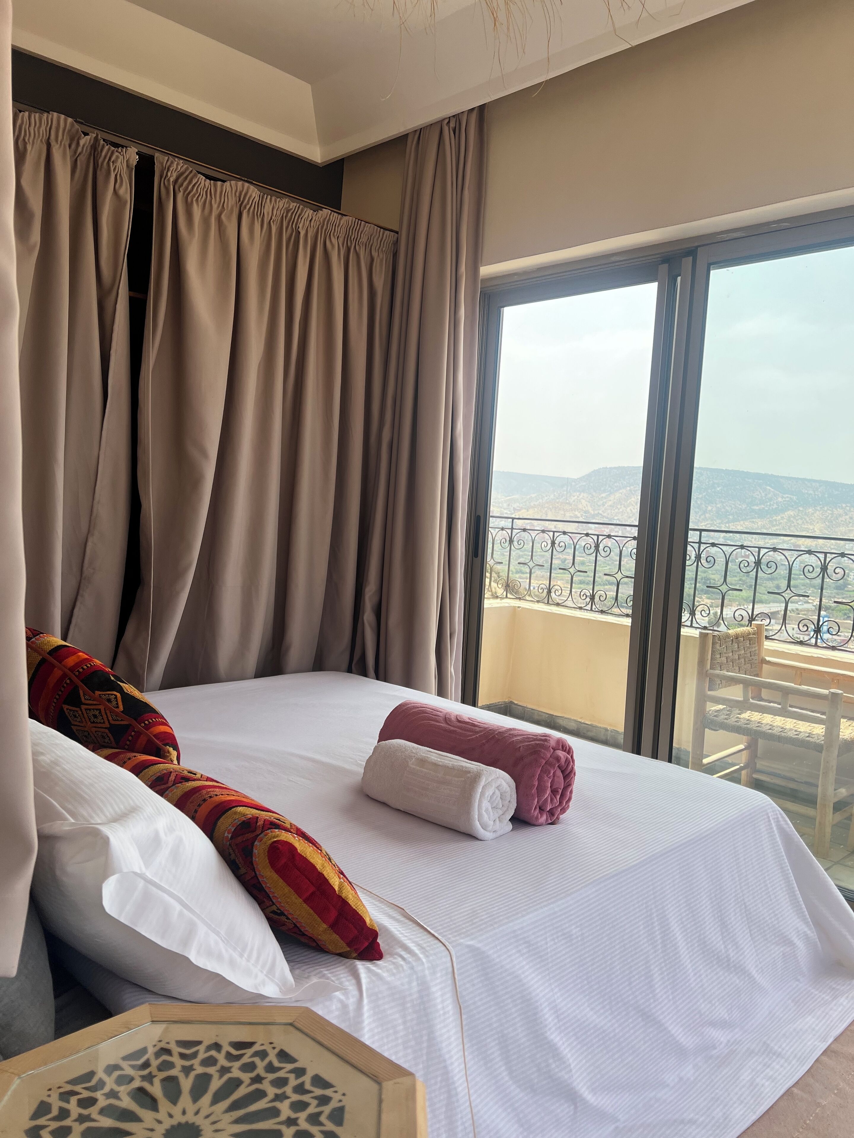 A hotel room with a large bed, beige curtains, a balcony with a view of the mountains, and a decorative wooden panel.