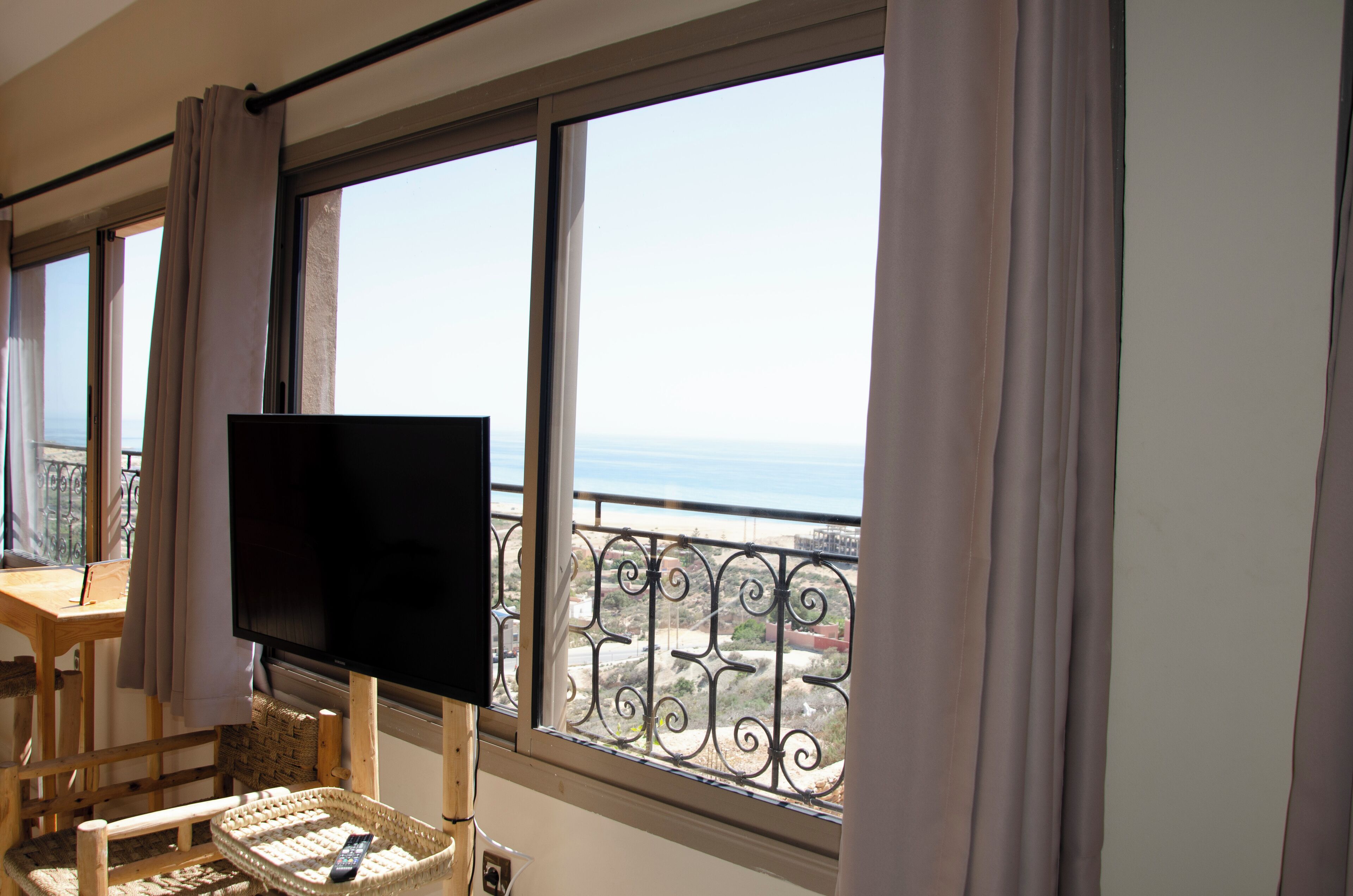 Una habitación con televisión sobre un mueble bajo, una silla, una mesita y vistas al mar a través de un balcón con barandilla de hierro.