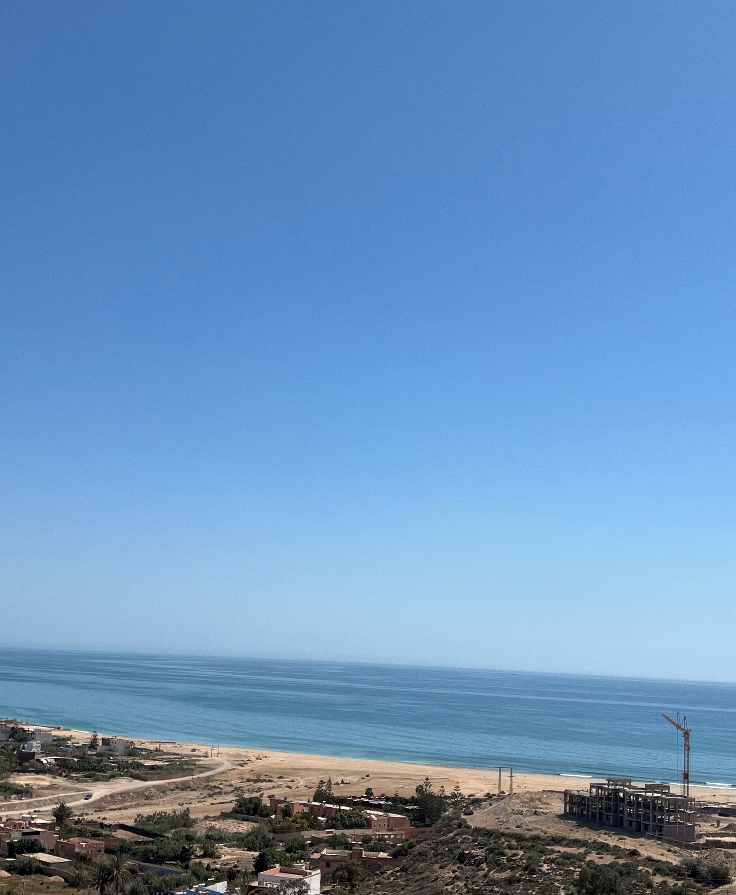 A coastal view with a clear sky, sandy beach, and a construction site with a crane.