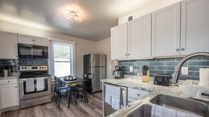 Una cocina moderna con armarios blancos, un refrigerador de acero inoxidable, un microondas, un horno y una cafetera. Hay una mesa de comedor con sillas y una ventana con persianas.