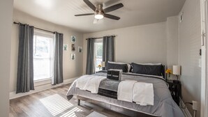 Un dormitorio con cama, ventilador de techo y dos ventanas con cortinas.