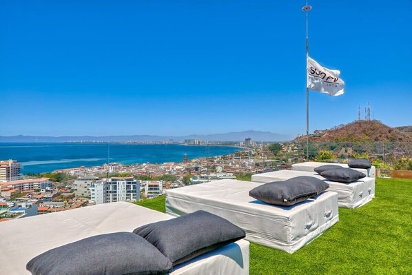 A rooftop terrace with several mattresses and pillows, overlooking a coastal cityscape with a flag in the background.