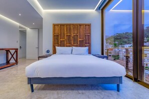 A modern bedroom with a large bed, a wooden headboard, a bedside table with a lamp, and a view of a cityscape through large windows.