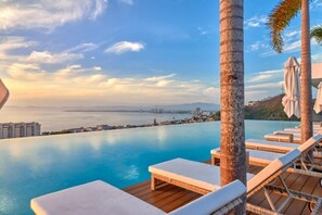 An infinity pool with lounge chairs and umbrellas, overlooking a coastal cityscape.