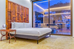 Ein Schlafzimmer mit einem großen Bett, einem hölzernen Kopfteil, einem kleinen Tisch und einem großen Fenster mit Blick auf die Stadt.